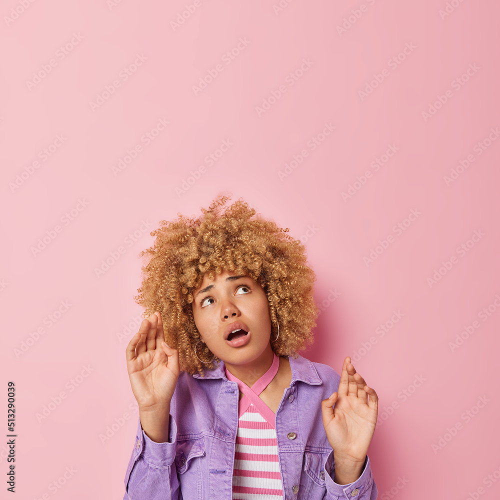 Scred woman keeps hands like something falling from above tries to protect herself from danger focused overhead wears purple jacket isolated over pink background empty space for your advert.