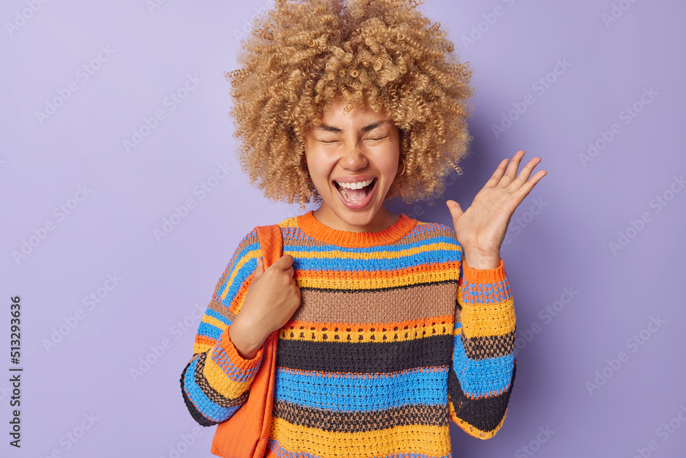 Big surprise concept. Overjoyed curly haired young woman exclaims with excitement reacts emotionally to good news carries bag wears casual colorful knitted jumper isolated ovr purple background
