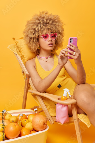 Beautiful curly haired woman addicted to modern technologies scrolls newsfeed via smartphone has lazy day spends summer vacations at beach wears pink sunglasses and swimsuit. Best holiday ever photo