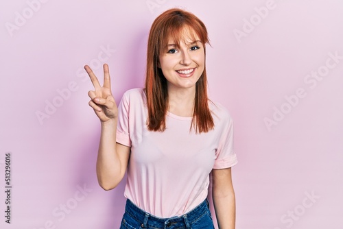 Redhead young woman wearing casual pink t shirt showing and pointing up with fingers number two while smiling confident and happy.