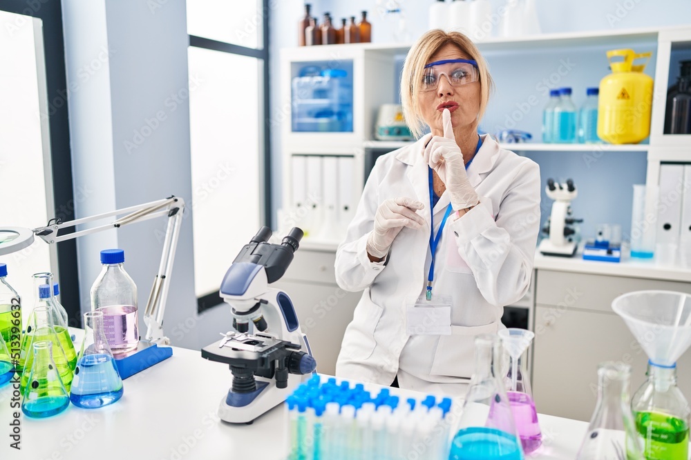 Middle age blonde woman working at scientist laboratory asking to be quiet with finger on lips. silence and secret concept. Middle age blonde woman working at scientist laboratory asking to be quiet with finger on lips. silence and secret concept.