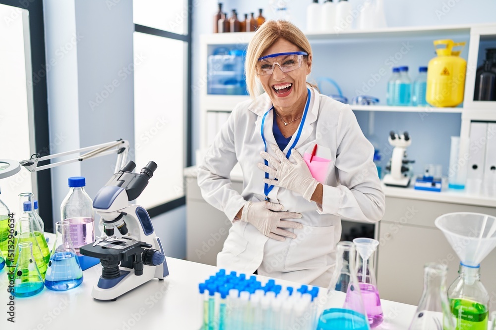Middle age blonde woman working at scientist laboratory smiling and laughing hard out loud because funny crazy joke with hands on body. Middle age blonde woman working at scientist laboratory smiling and laughing hard out loud because funny crazy joke with hands on body.