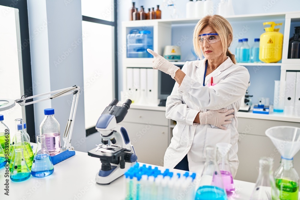 Middle age blonde woman working at scientist laboratory pointing with hand finger to the side showing advertisement, serious and calm face Middle age blonde woman working at scientist laboratory pointing with hand finger to the side showing advertisement, serious and calm face