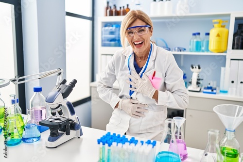 Middle age blonde woman working at scientist laboratory smiling and laughing hard out loud because funny crazy joke with hands on body.
