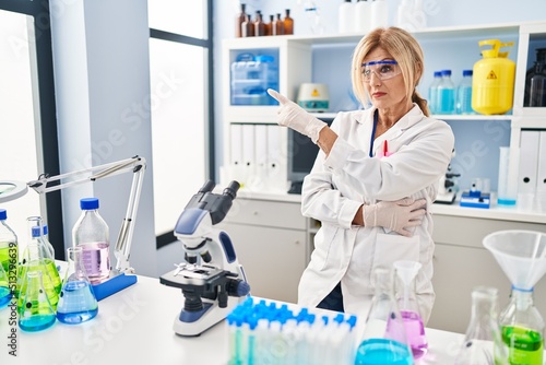 Middle age blonde woman working at scientist laboratory pointing with hand finger to the side showing advertisement  serious and calm face