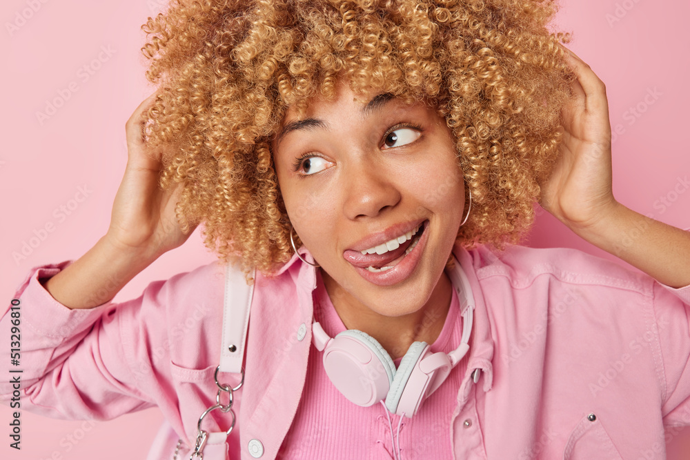 Dreamy teenage girl sticks out tongue looks away pensively keeps hands on her curly bushy hair wears fashionable jacket and headphones around neck for listening music isolated over pink background. Dreamy teenage girl sticks out tongue looks away pensively keeps hands on her curly bushy hair wears fashionable jacket and headphones around neck for listening music isolated over pink background.