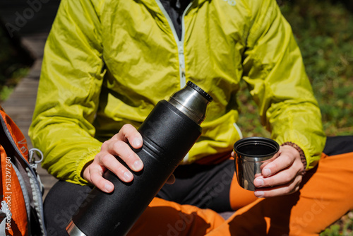 The guy holds in his hand a thermos with hot tea, dishes for a hike, lunch in nature, a thermo glass with coffee, camping equipment utensils.