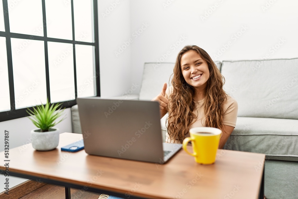 Beautiful hispanic woman using computer laptop at home smiling happy and positive, thumb up doing excellent and approval sign Beautiful hispanic woman using computer laptop at home smiling happy and positive, thumb up doing excellent and approval sign