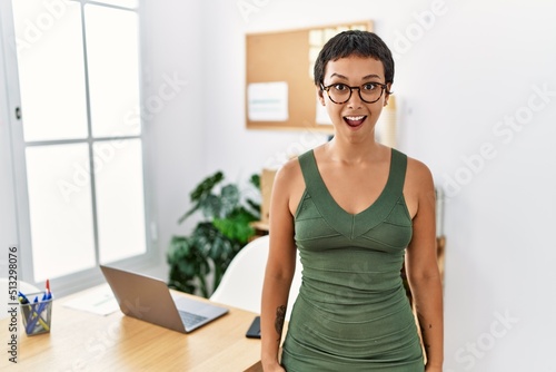Young hispanic woman with short hair working at the office scared and amazed with open mouth for surprise, disbelief face