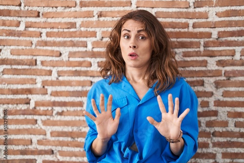 Beautiful brunette woman standing over bricks wall moving away hands palms showing refusal and denial with afraid and disgusting expression. stop and forbidden.