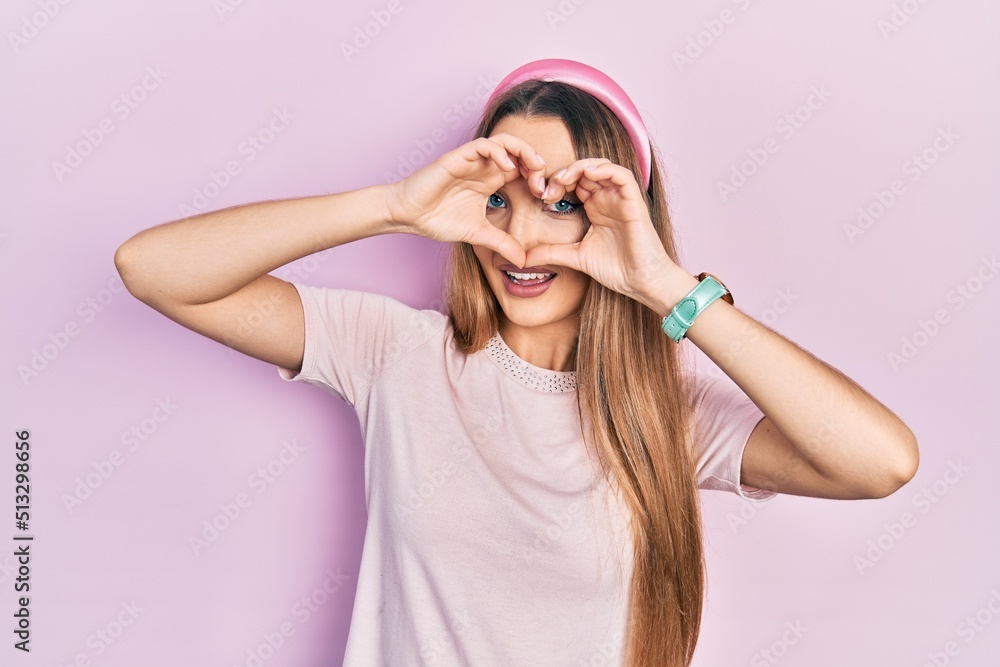 Young blonde girl wearing casual clothes doing heart shape with hand and fingers smiling looking through sign
