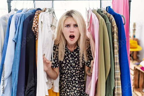 Young blonde woman searching clothes on clothing rack afraid and shocked with surprise and amazed expression, fear and excited face.