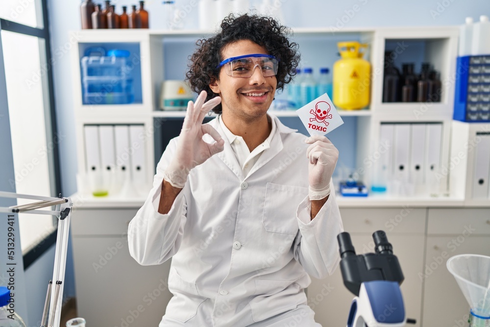 Hispanic man with curly hair working at scientist laboratory holding toxic banner doing ok sign with fingers, smiling friendly gesturing excellent symbol Hispanic man with curly hair working at scientist laboratory holding toxic banner doing ok sign with fingers, smiling friendly gesturing excellent symbol