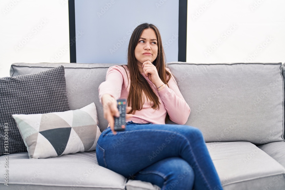 Young brunette woman holding television remote control serious face thinking about question with hand on chin, thoughtful about confusing idea Young brunette woman holding television remote control serious face thinking about question with hand on chin, thoughtful about confusing idea