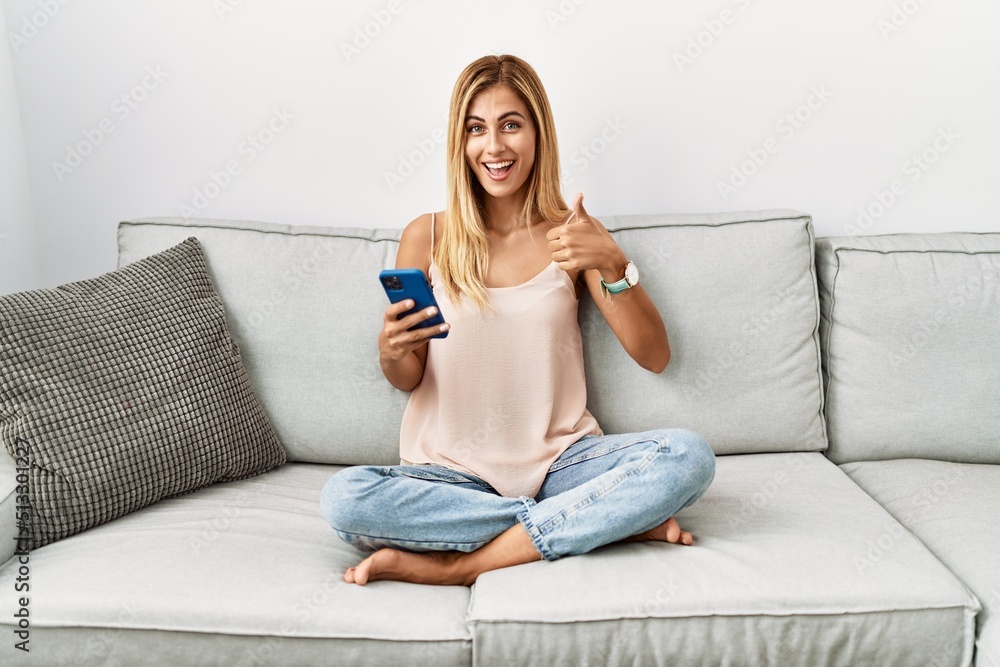 Blonde beautiful young woman sitting on the sofa at home using smartphone doing happy thumbs up gesture with hand. approving expression looking at the camera showing success. Blonde beautiful young woman sitting on the sofa at home using smartphone doing happy thumbs up gesture with hand. approving expression looking at the camera showing success.