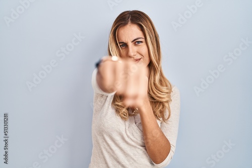 Young blonde woman standing over isolated background punching fist to fight, aggressive and angry attack, threat and violence