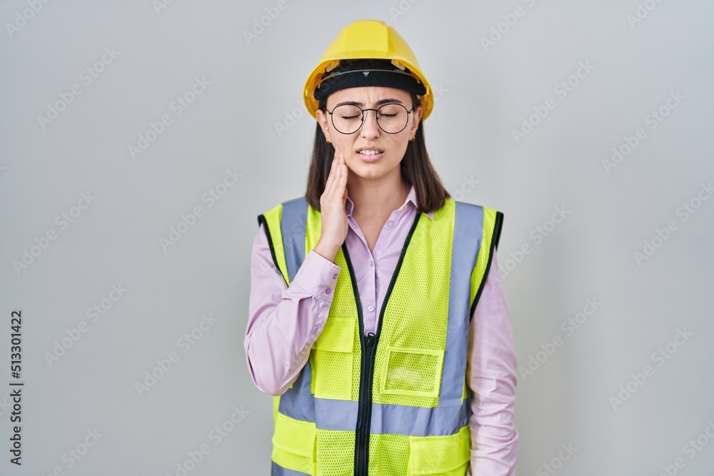 Hispanic girl wearing builder uniform and hardhat touching mouth with hand with painful expression because of toothache or dental illness on teeth. dentist Hispanic girl wearing builder uniform and hardhat touching mouth with hand with painful expression because of toothache or dental illness on teeth. dentist