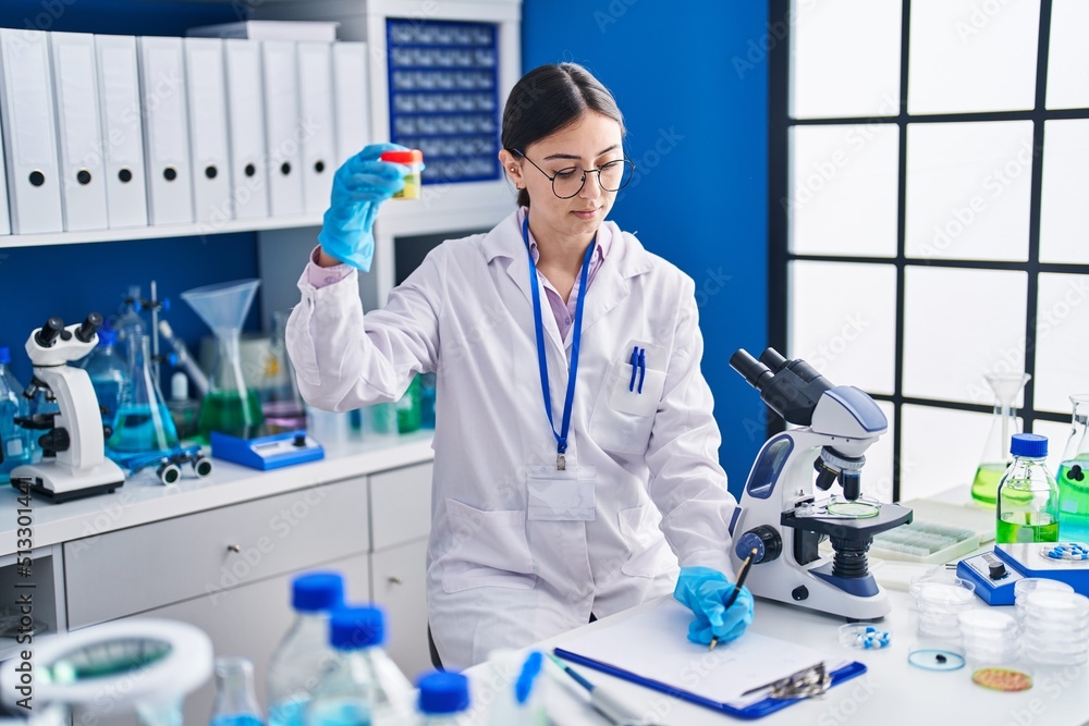 Young hispanic woman scientist writing on document holding urine test tube at laboratory
