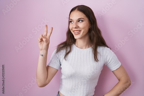 Young hispanic girl standing over pink background smiling looking to the camera showing fingers doing victory sign. number two.