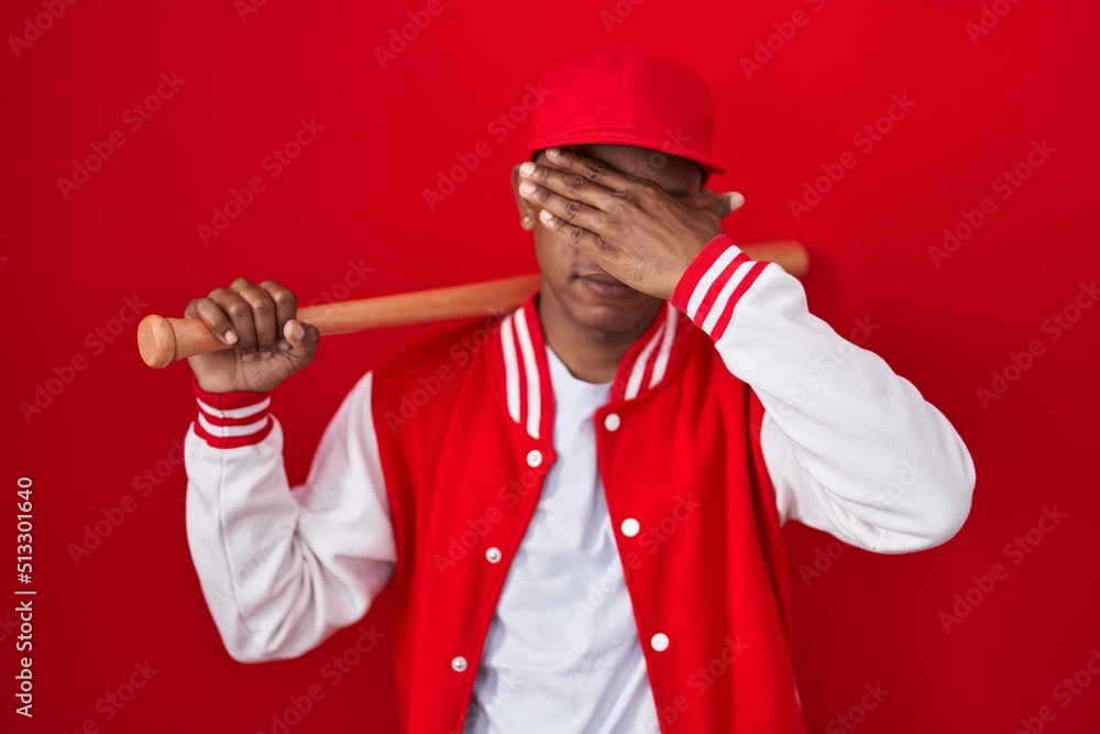 Young hispanic man playing baseball holding bat covering eyes with hand, looking serious and sad. sightless, hiding and rejection concept Young hispanic man playing baseball holding bat covering eyes with hand, looking serious and sad. sightless, hiding and rejection concept