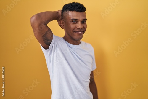 Young hispanic man standing over yellow background smiling confident touching hair with hand up gesture, posing attractive and fashionable