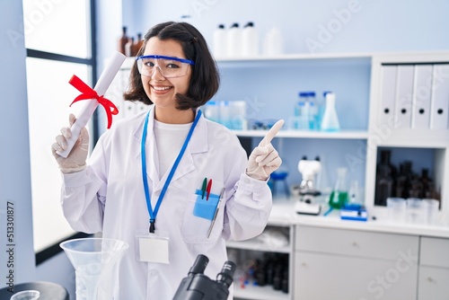 Young hispanic woman working at scientist laboratory holding degree smiling happy pointing with hand and finger to the side