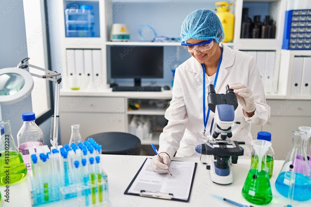 Fototapeta premium Young latin woman wearing scientist uniform using microscope at laboratory