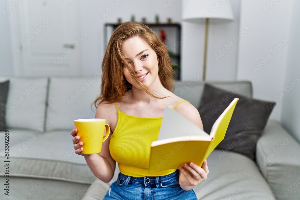 Young redhead girl reading book drinking coffee at home