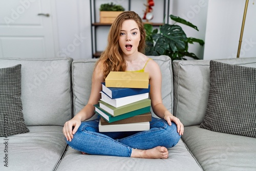 Young caucasian woman sitting on the sofa with books at home afraid and shocked with surprise expression, fear and excited face.