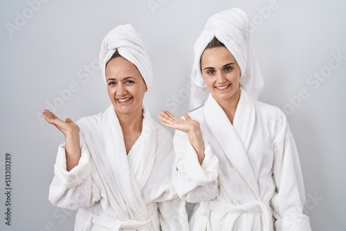 Middle age woman and daughter wearing white bathrobe and towel smiling cheerful presenting and pointing with palm of hand looking at the camera.