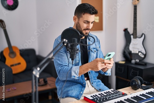 Young hispanic man artist smiling confident using smartphone at music studio