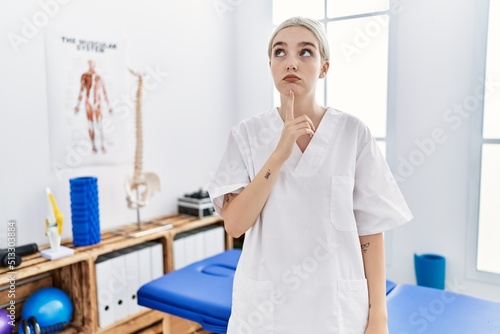 Young caucasian woman working at pain recovery clinic thinking concentrated about doubt with finger on chin and looking up wondering