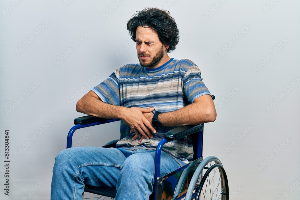 Handsome hispanic man sitting on wheelchair with hand on stomach because indigestion, painful illness feeling unwell. ache concept. Handsome hispanic man sitting on wheelchair with hand on stomach because indigestion, painful illness feeling unwell. ache concept.