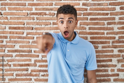 Brazilian young man standing over brick wall pointing with finger surprised ahead, open mouth amazed expression, something on the front