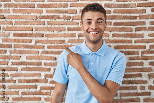 Brazilian young man standing over brick wall cheerful with a smile on face pointing with hand and finger up to the side with happy and natural expression