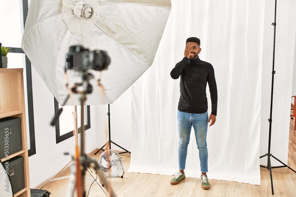 Young hispanic man with beard posing as model at photography studio covering one eye with hand, confident smile on face and surprise emotion.