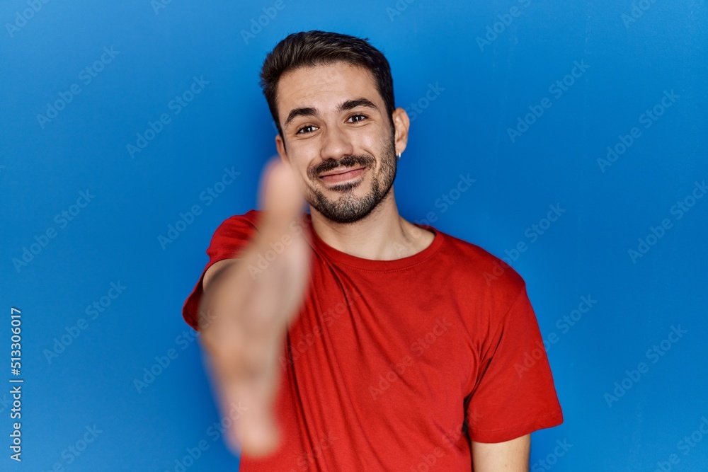 Young hispanic man with beard wearing red t shirt over blue background smiling friendly offering handshake as greeting and welcoming. successful business. Young hispanic man with beard wearing red t shirt over blue background smiling friendly offering handshake as greeting and welcoming. successful business.