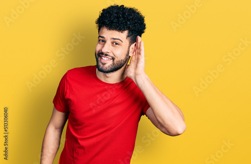 Young arab man with beard wearing casual red t shirt smiling with hand over ear listening an hearing to rumor or gossip. deafness concept.