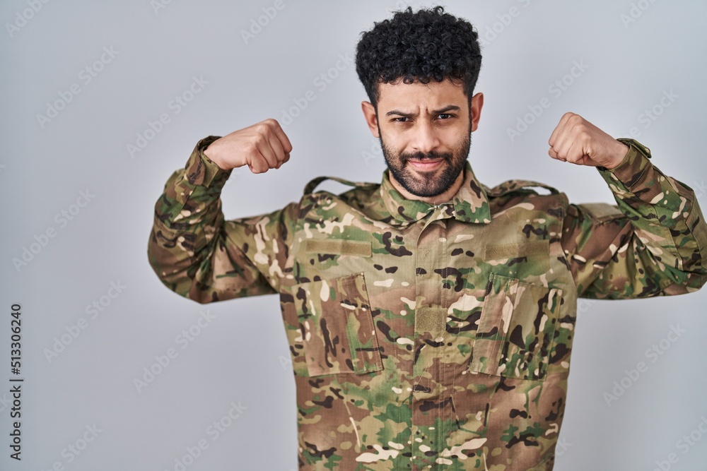 Arab man wearing camouflage army uniform showing arms muscles smiling proud. fitness concept.