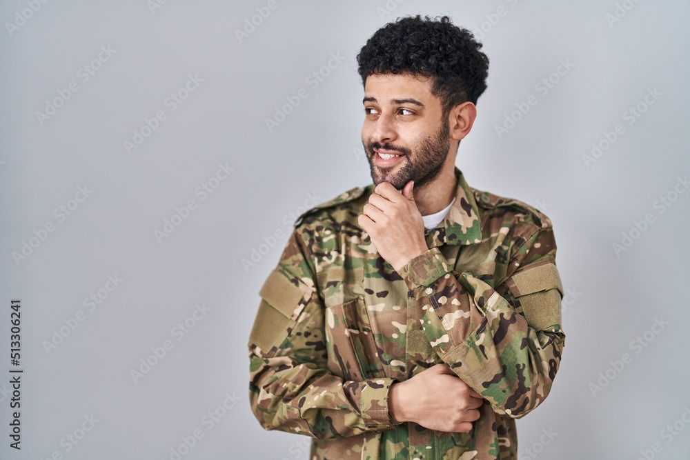 Arab man wearing camouflage army uniform with hand on chin thinking about question, pensive expression. smiling and thoughtful face. doubt concept.
