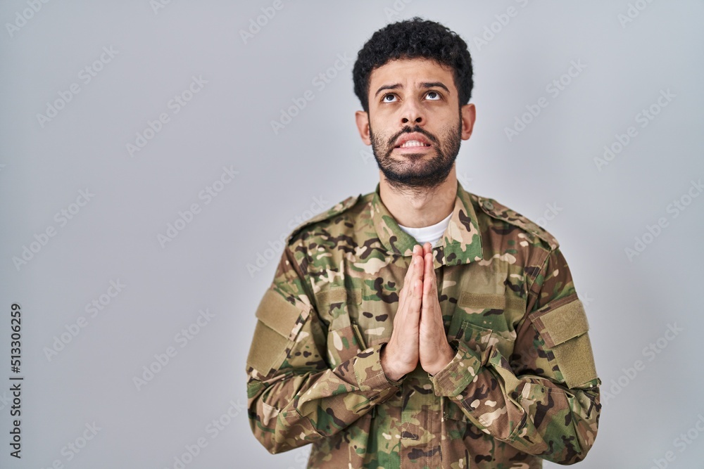 Arab man wearing camouflage army uniform begging and praying with hands together with hope expression on face very emotional and worried. begging.