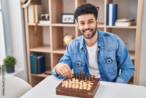 Young arab man playing chess sitting on table at home