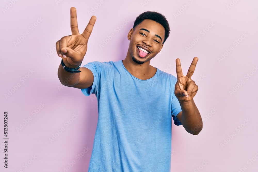 Young african american man wearing casual blue t shirt smiling with tongue out showing fingers of both hands doing victory sign. number two. Young african american man wearing casual blue t shirt smiling with tongue out showing fingers of both hands doing victory sign. number two.