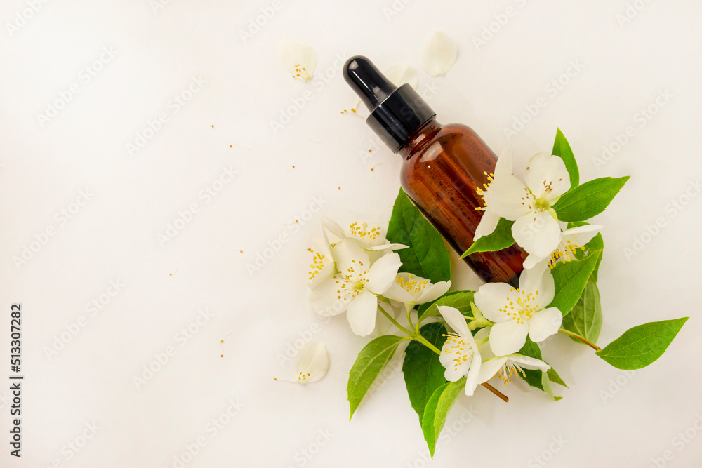 Cosmetic brown glass bottle with a dispenser on a white background. White flowers with green leaves of an ornamental shrub. Philadelphus or Jasminum. Cosmetic brown glass bottle with a dispenser on a white background. White flowers with green leaves of an ornamental shrub. Philadelphus or Jasminum.