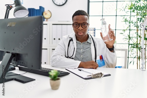 Young african doctor man holding hand sanitizer gel at the clinic thinking attitude and sober expression looking self confident