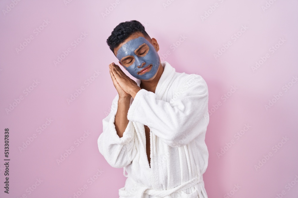 Young hispanic man wearing beauty face mask and bath robe sleeping tired dreaming and posing with hands together while smiling with closed eyes. Young hispanic man wearing beauty face mask and bath robe sleeping tired dreaming and posing with hands together while smiling with closed eyes.