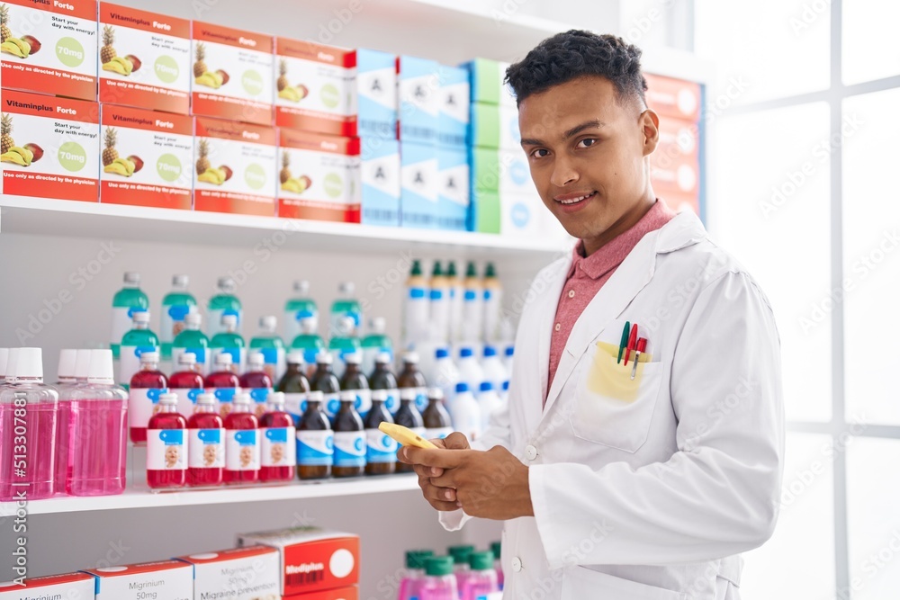 Young latin man pharmacist smiling confident using smartphone at pharmacy