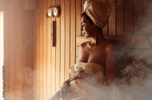 Young woman relaxing and sweating in hot sauna wrapped in towel. Girl In Sauna. Interior of Finnish sauna, classic wooden sauna with hot steam. Russian bathroom. Relax in hot sauna with steam photo