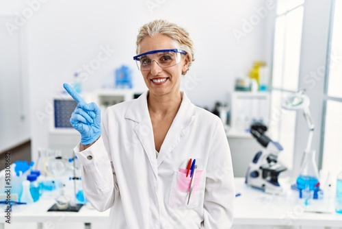 Middle age blonde woman working at scientist laboratory with a big smile on face, pointing with hand and finger to the side looking at the camera.