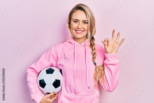 Beautiful young blonde woman holding soccer ball doing ok sign with fingers  smiling friendly gesturing excellent symbol